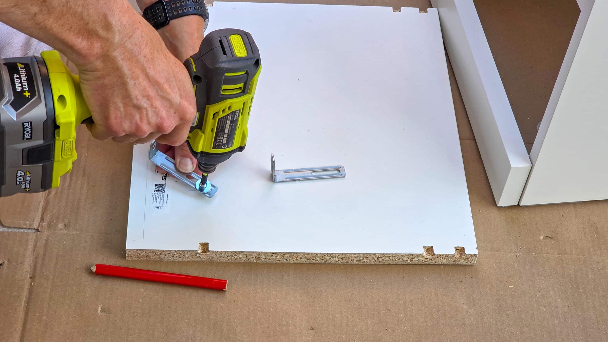 A person uses a cordless power drill to attach a metal bracket to a white chipboard panel, with a red pencil and another bracket nearby, assembling furniture on a cardboard surface.