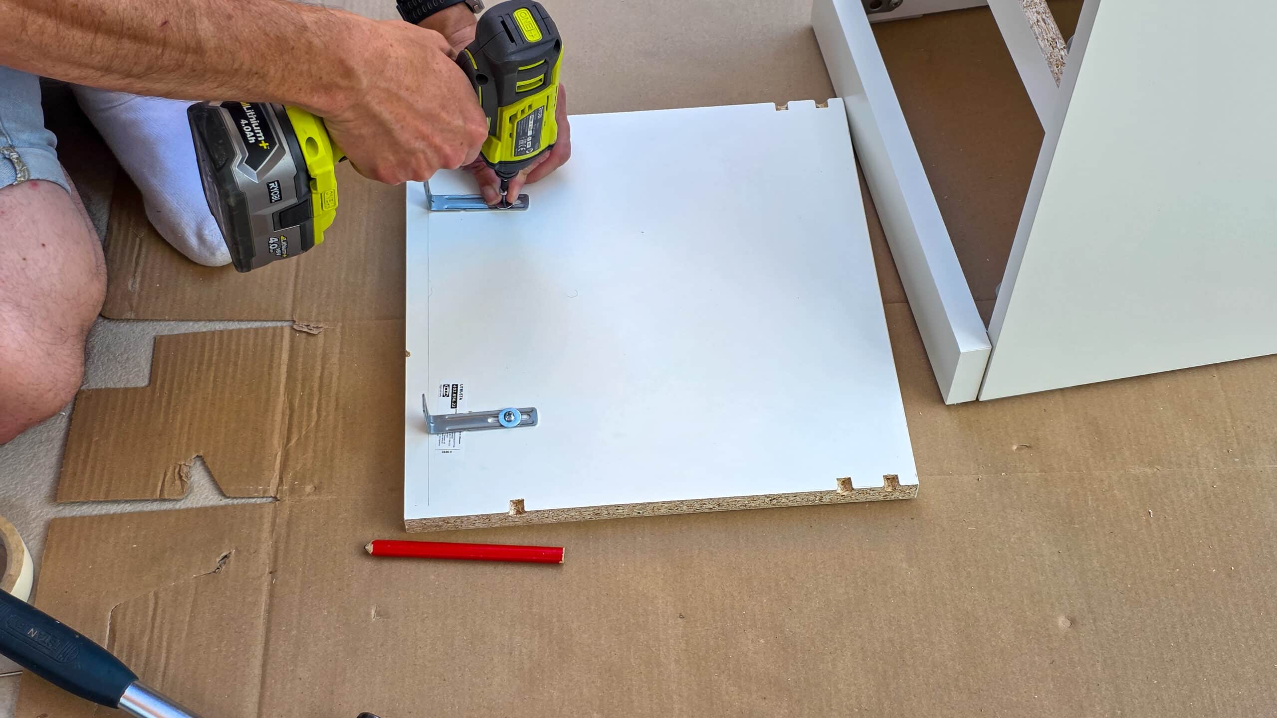 A person uses a cordless drill to attach metal brackets to a white wooden board, with a red pencil and cupboard parts nearby on a cardboard-covered surface.