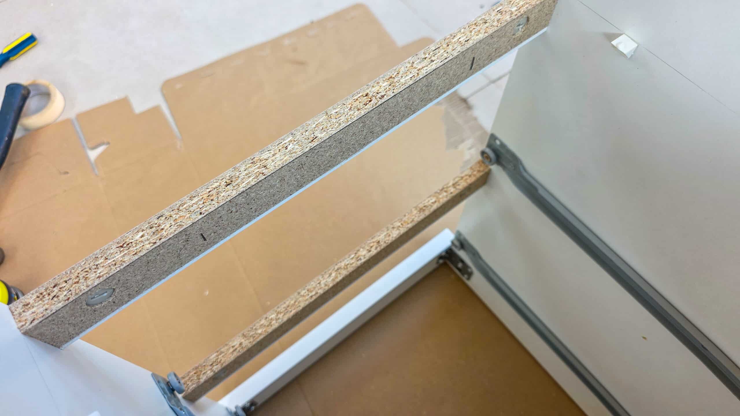 Close-up of a partially assembled drawer or cabinet made of chipboard, showing unfinished edges and metal drawer runners on the sides, with tools and cardboard on the floor in the background.