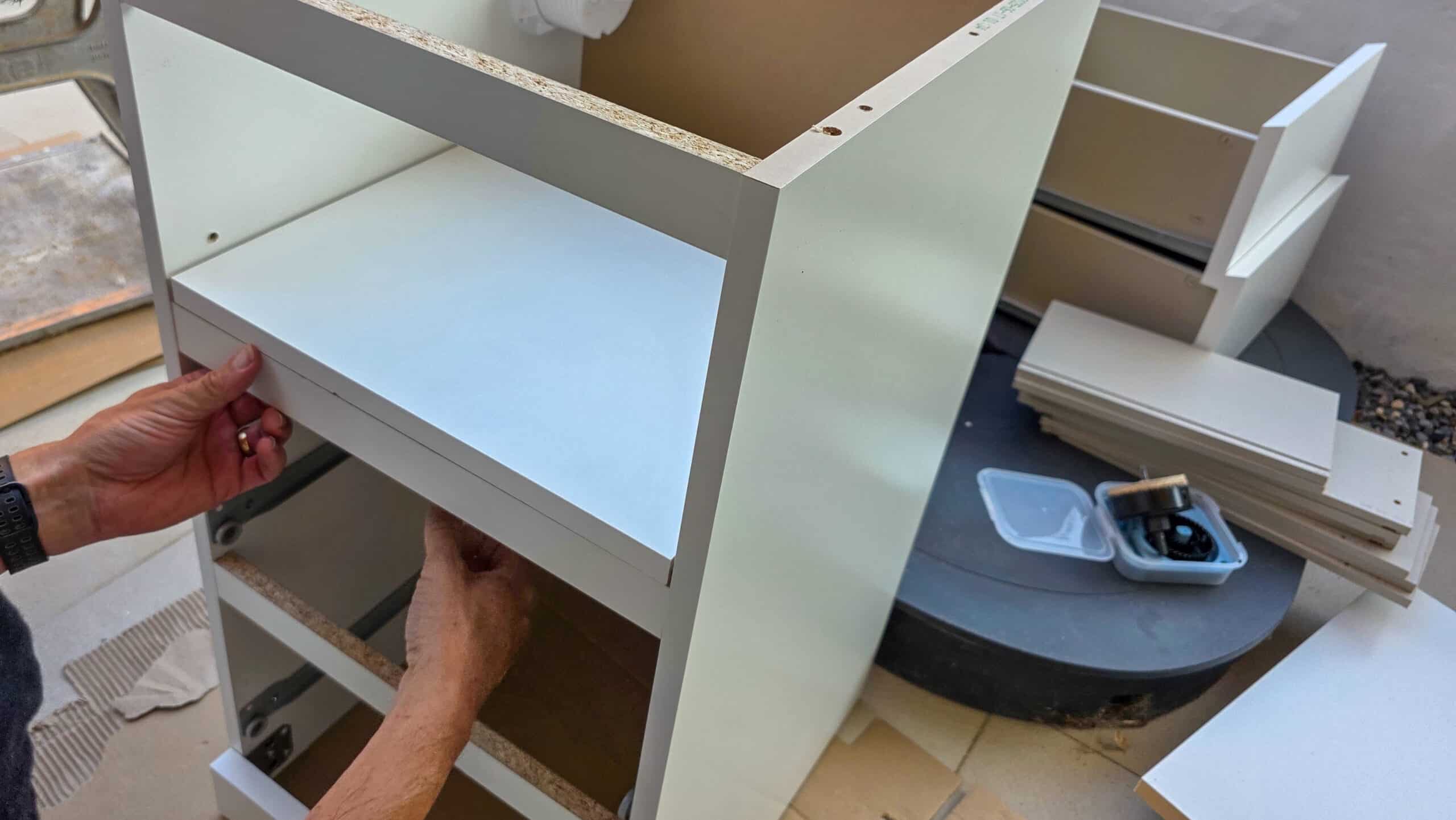 A person assembling a white wooden cupboard, positioning a shelf, with drawers and other parts scattered nearby, along with a small container of screws and fittings.
