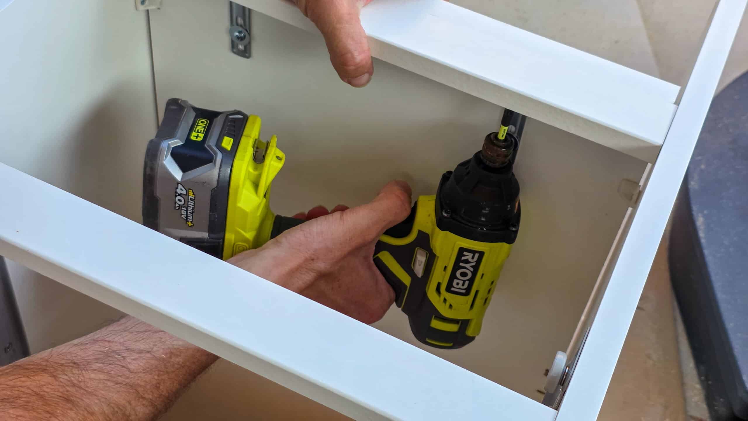 A person uses a yellow and black Ryobi cordless drill to assemble a white cupboard, holding the drill inside the frame while securing a screw.