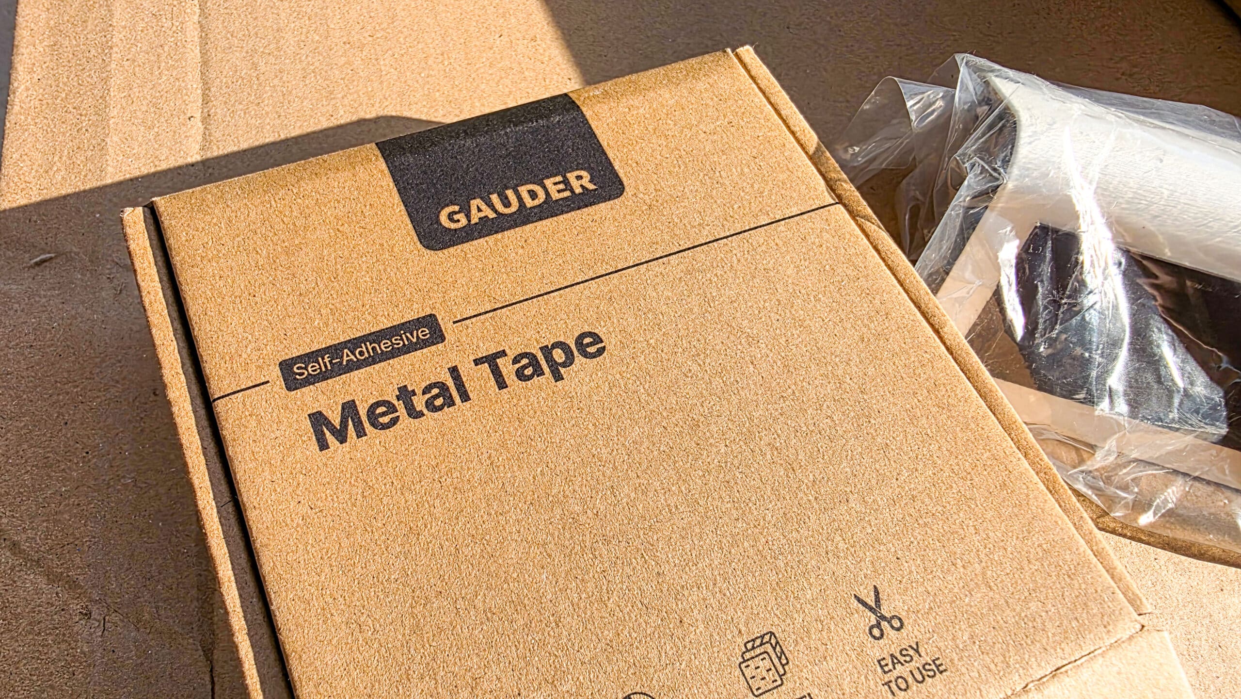 A close-up of a cardboard box labelled GAUDER Self-Adhesive Metal Tape with icons showing product features, next to a partially visible tape dispenser wrapped in plastic.