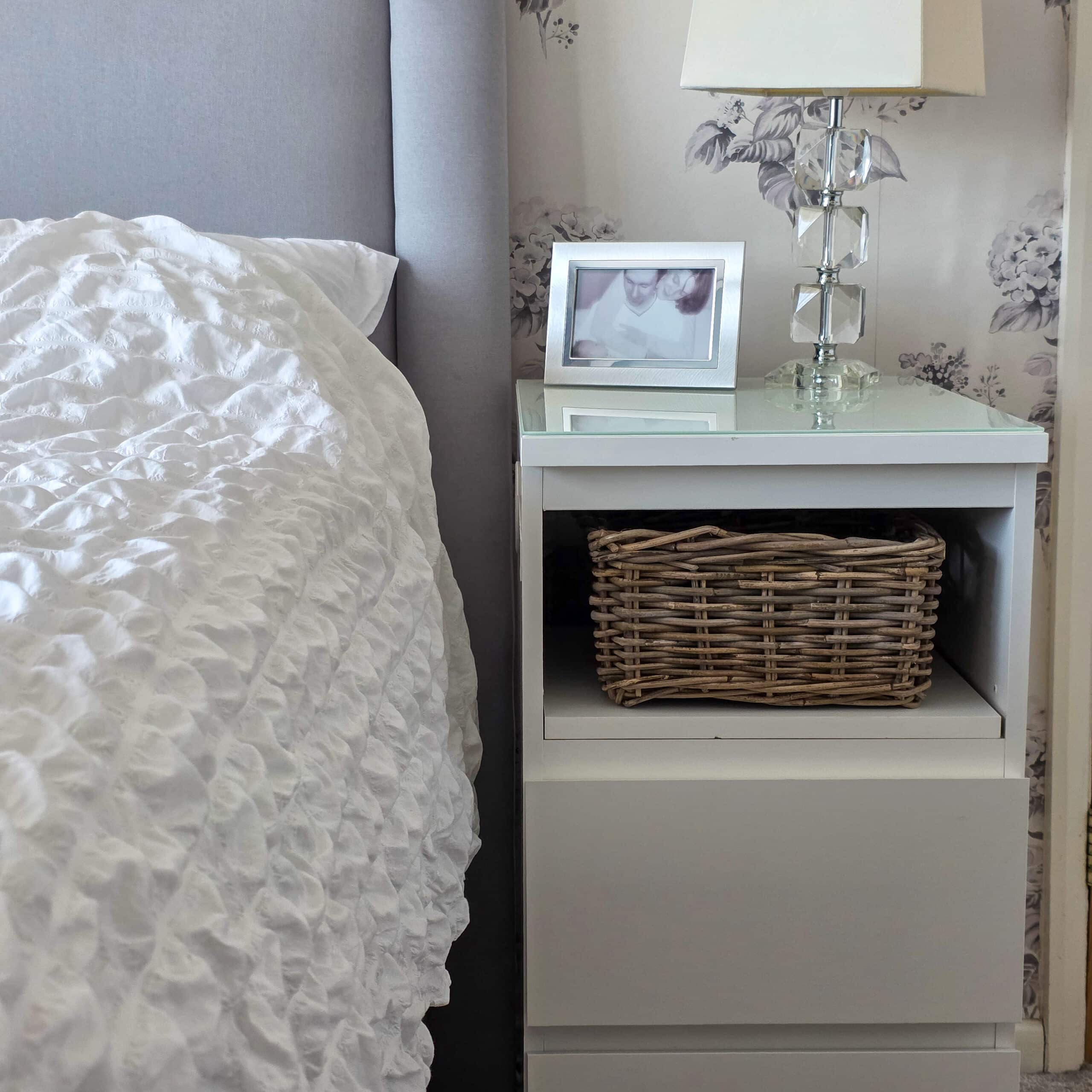 IKEA Malm Bedside Table Hack. A white bedside table with a wicker basket on a shelf, a glass lamp, and a framed photo on top, next to a bed with a textured white duvet. The wall has light floral-patterned wallpaper.