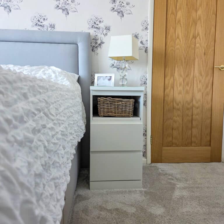 IKEA Malm Bedside Table Hack With Phone Charging - A modern white bedside table next to a bed with a textured white duvet, featuring a wicker basket, a framed photo, and a glass lamp, set against floral wallpaper and a closed wooden door.