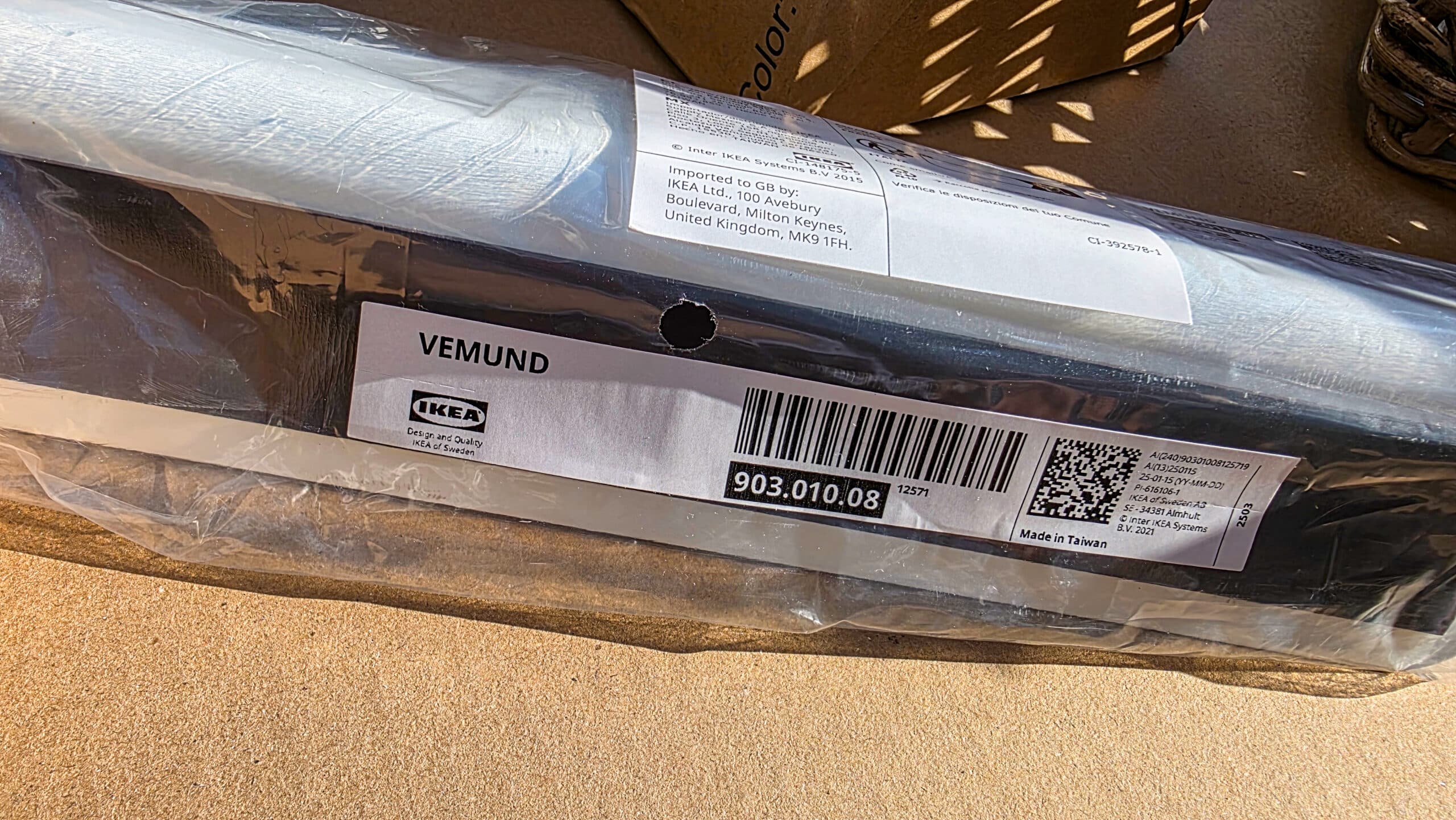 A close-up photo of an IKEA VEMUND product in its packaging, showing the label with the item number 903.010.08, barcode, and manufacturing details, placed on a brown surface.
