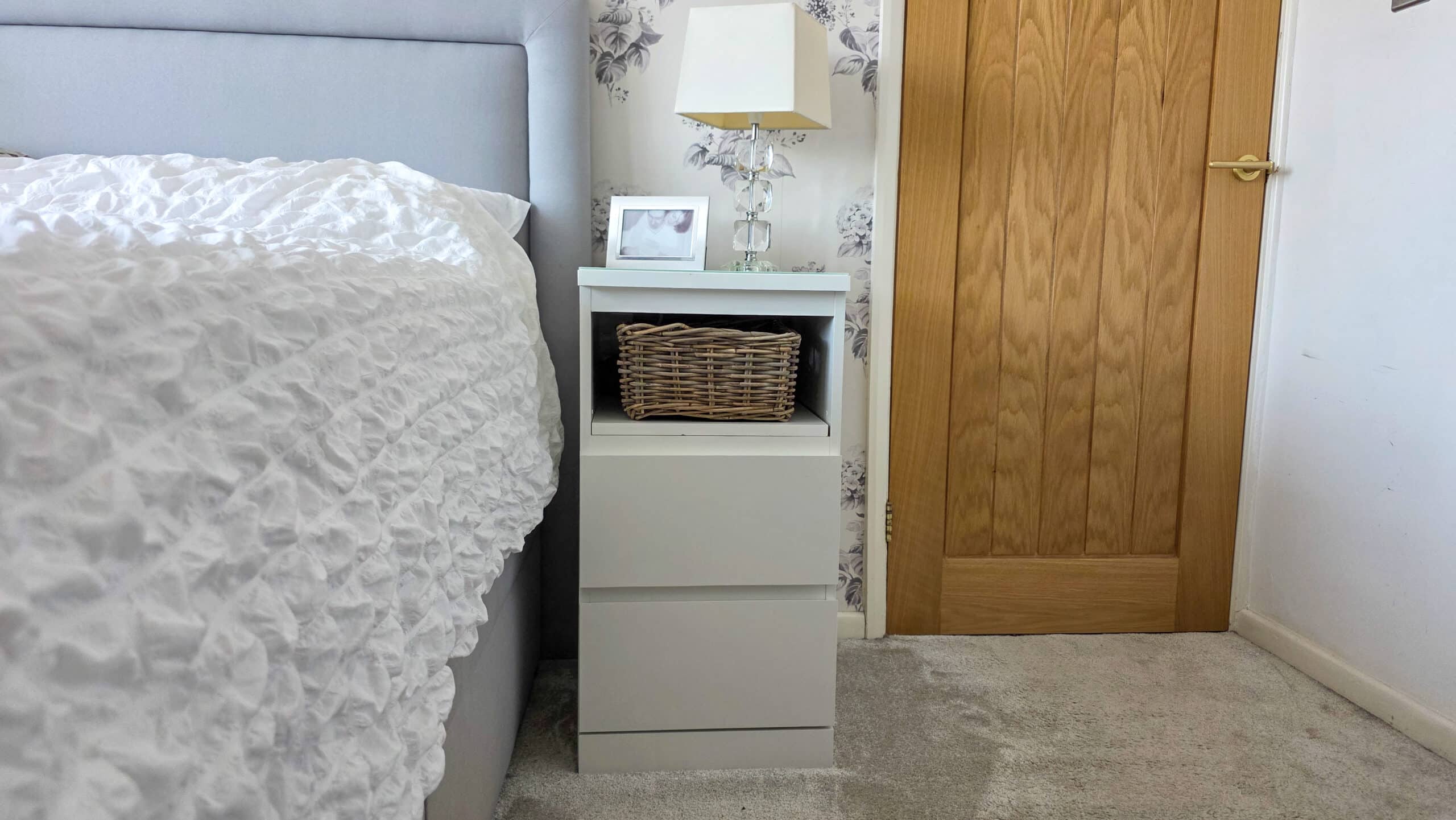 IKEA Malm Bedside Table Hack With Phone Charging. A white bedside table with two drawers, a wicker basket, a small framed photo, and a lamp sits next to a bed with a textured white duvet, near a wooden door on light carpeted flooring.