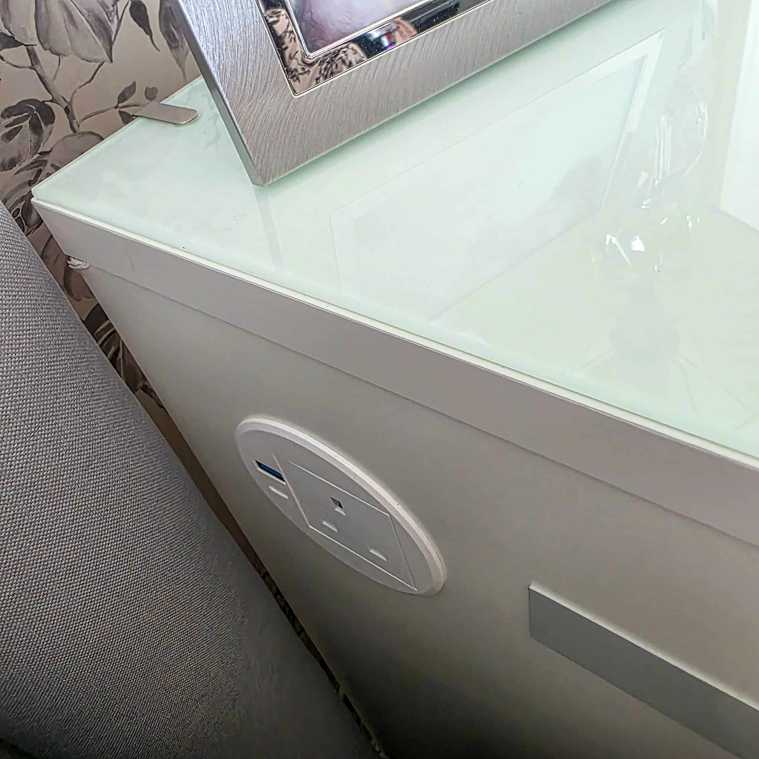 IKEA Malm Bedside Table Hack With Phone Charging. Close-up of a white power outlet with a UK plug socket and USB ports on the side of a white bedside table next to a grey upholstered surface and floral wallpaper. A photo frame and lamp are on the glass table top.
