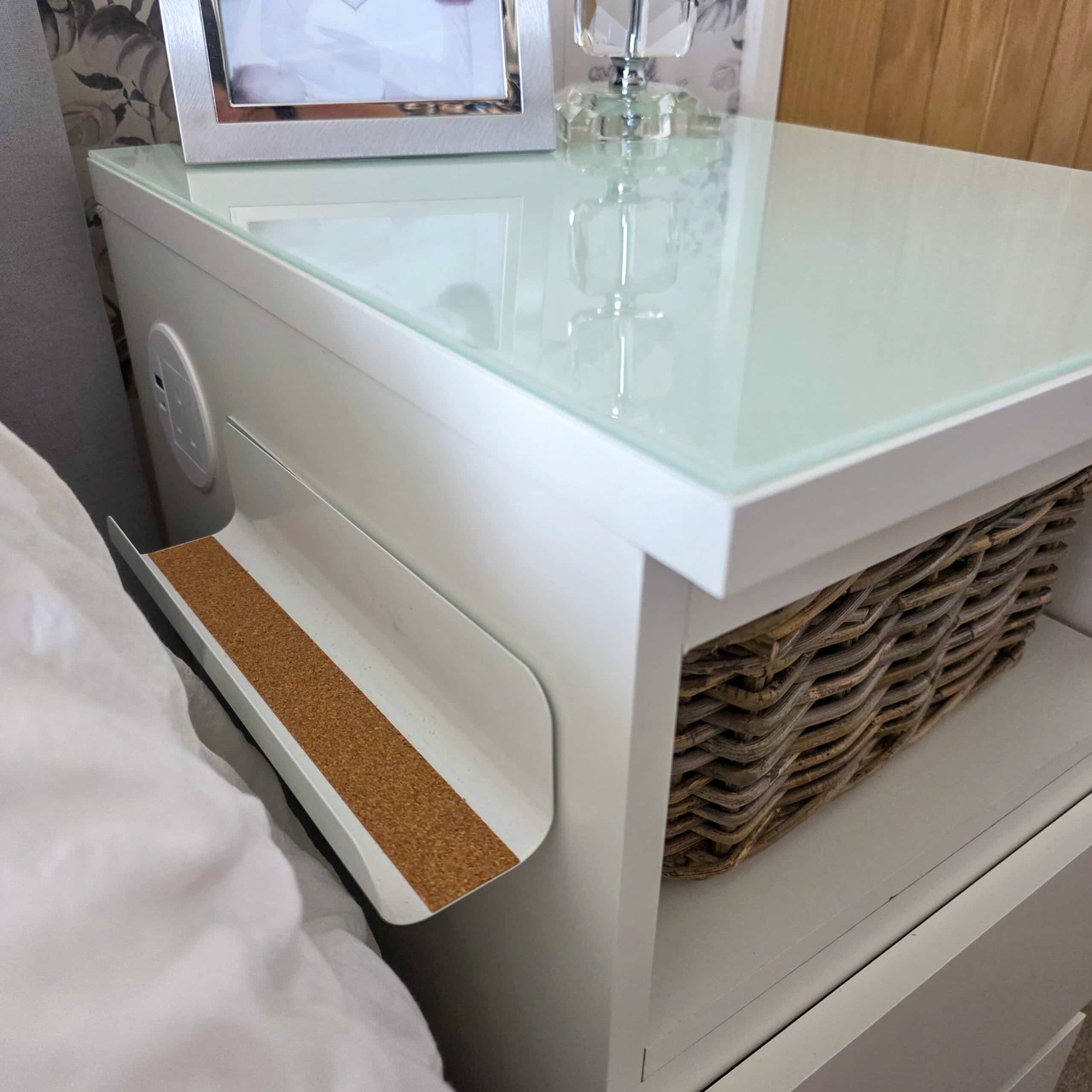 IKEA Malm Bedside Table Hack With Phone Charging. A white bedside table with a glass top, a framed photo, a lamp, a built-in power socket, a cork-lined shelf, and a woven basket on a lower shelf. Part of a bed and a wooden door are visible.