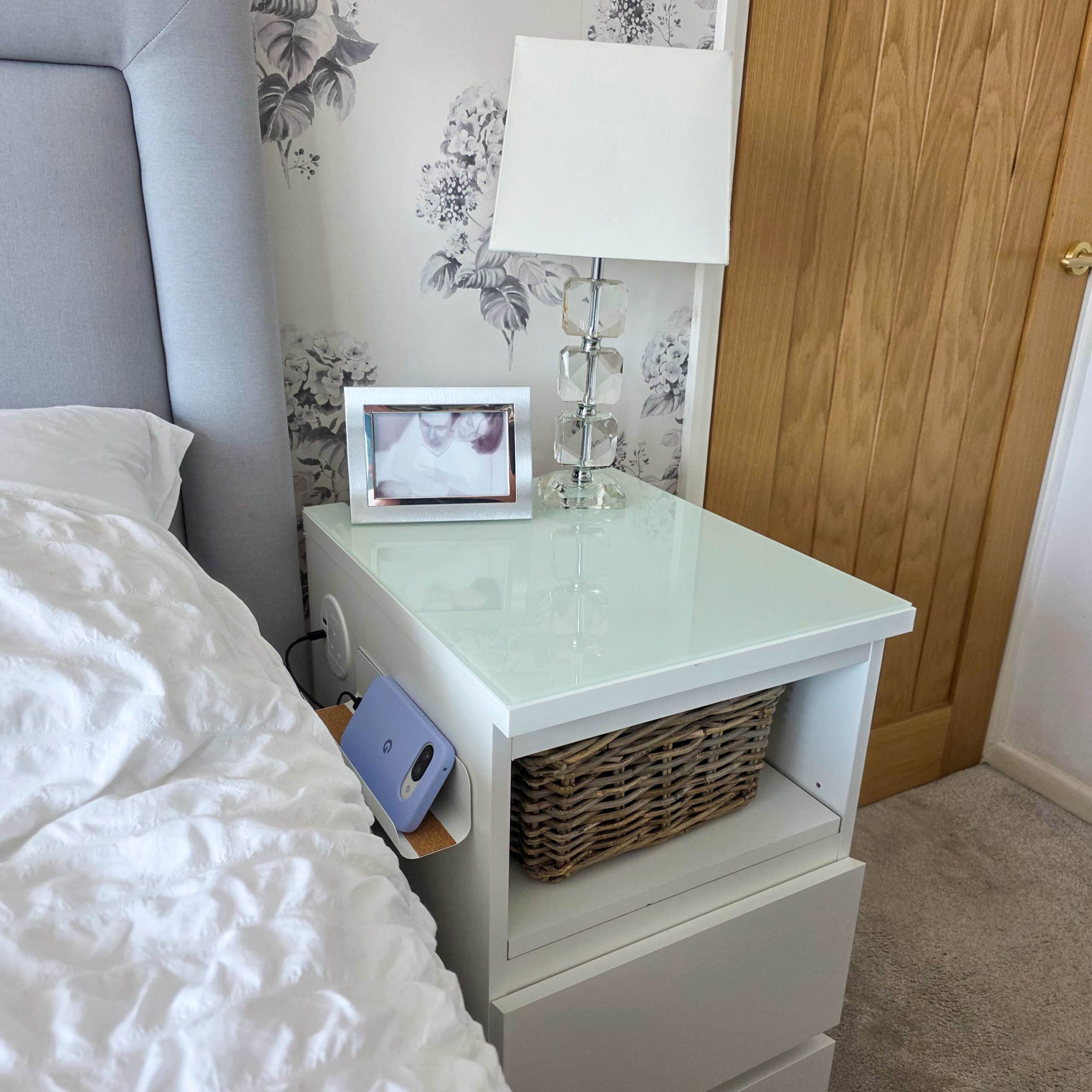 IKEA Malm Bedside Table Hack With Phone Charging. A white bedside table with a wicker basket, lamp, and framed photo on top stands next to a bed with white bedding. A wooden door is in the background, and a phone charges at the side of the table.