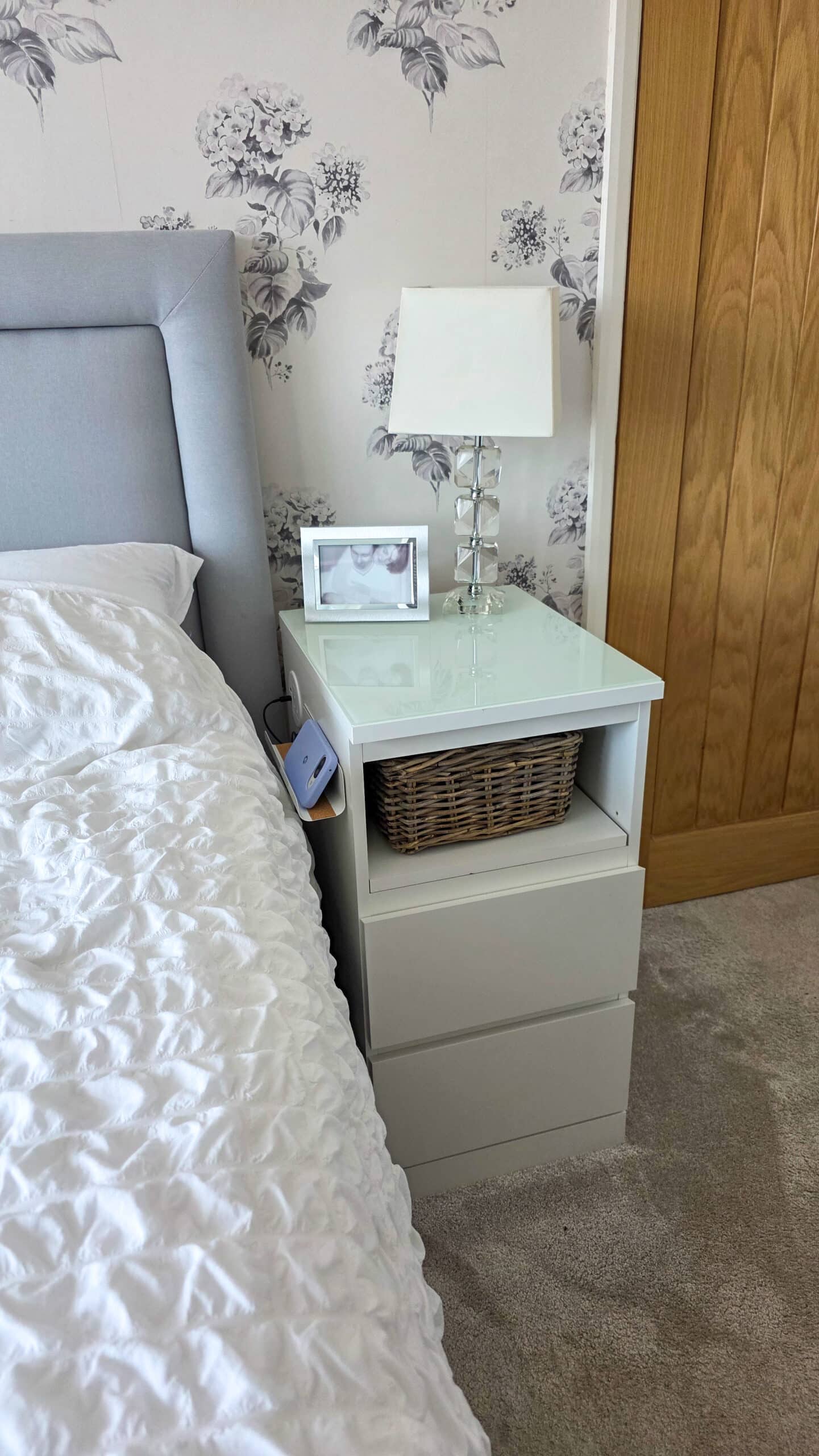 IKEA Malm Bedside Table Hack With Phone Charging. A neatly made bed with a white duvet is next to a white bedside table holding a lamp, a framed photo, and a wicker basket. The wall behind features floral wallpaper and a wooden door is to the right.