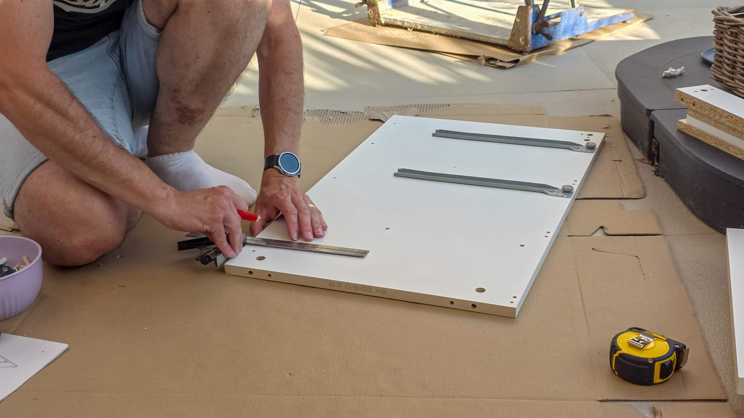 A person in shorts measures and marks a white wooden board with a ruler and pencil, sitting on cardboard. Tools and materials, including a tape measure, are nearby, indicating furniture assembly.