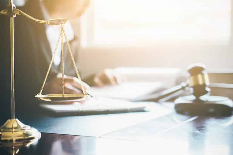 A brass balance scale and a wooden gavel on a desk, with a person writing in the background, suggest a legal or courtroom setting—perfect for illustrating the roles of Solicitor vs Independent Legal Adviser. Sunlight streams in through a nearby window.