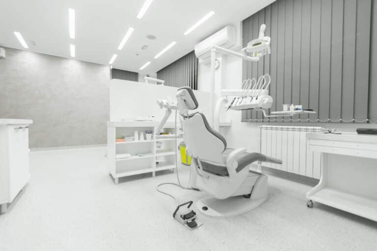 A modern, clean dental surgery room with a grey dental chair, overhead light, white cupboards, and equipment trays. Vertical grey blinds cover the windows in this brightly lit space designed to support personalised health plans.