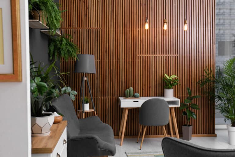 Modern home office with a feature wall of MDF wall cladding and wooden slats, grey chair, white desk with potted plants and cacti, grey floor lamp, and hanging lights, surrounded by green houseplants for a cosy, stylish look.