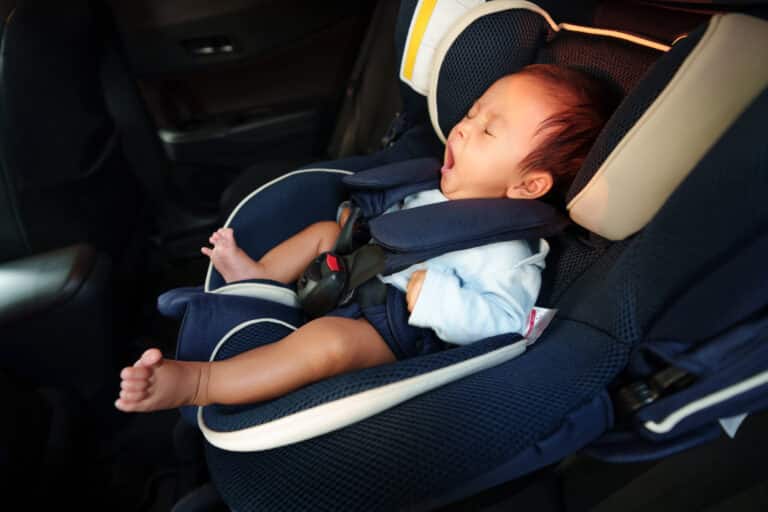A baby yawns while sitting in a rear-facing car seat, safely strapped in with a harness, embodying the comfort and security essential for the psychology of travel. The baby appears relaxed, with one leg stretched out inside the car.