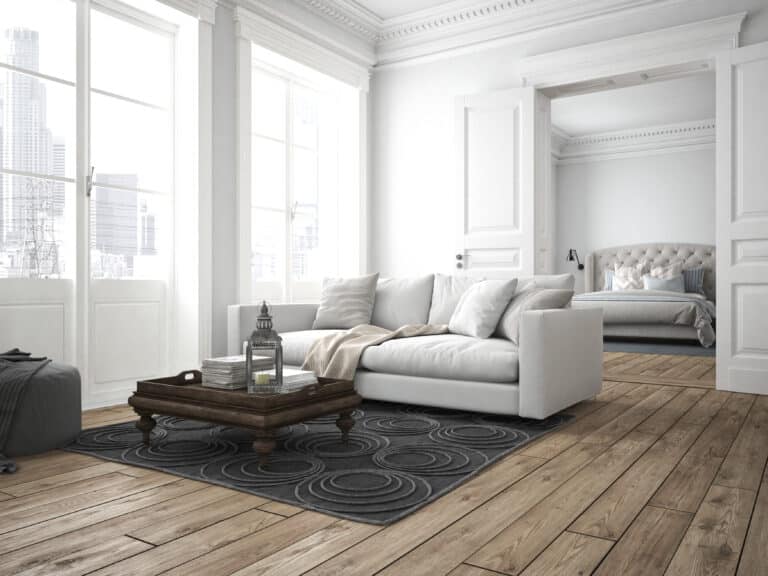Bright, modern living room with a white sofa, wooden coffee table, grey rug, and large windows designed for urban comfort. An open doorway leads to a bedroom with a tufted headboard and neutral bedding—choose an Architect Central London for style like this.