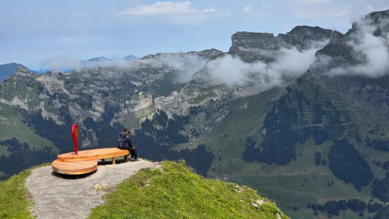 A person sits alone on a curved wooden bench atop a grassy mountain ledge, reflecting on Solicitor vs Independent Legal Adviser while overlooking a valley and rugged mountains partially covered by low clouds under a blue sky.