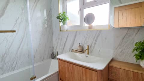 Modern small bathroom before and after: marble walls, bath with glass panel, wooden vanity with white basin, gold tap, plants on the windowsill and counter, and a mirror above the basin. Natural light shines through a frosted window.