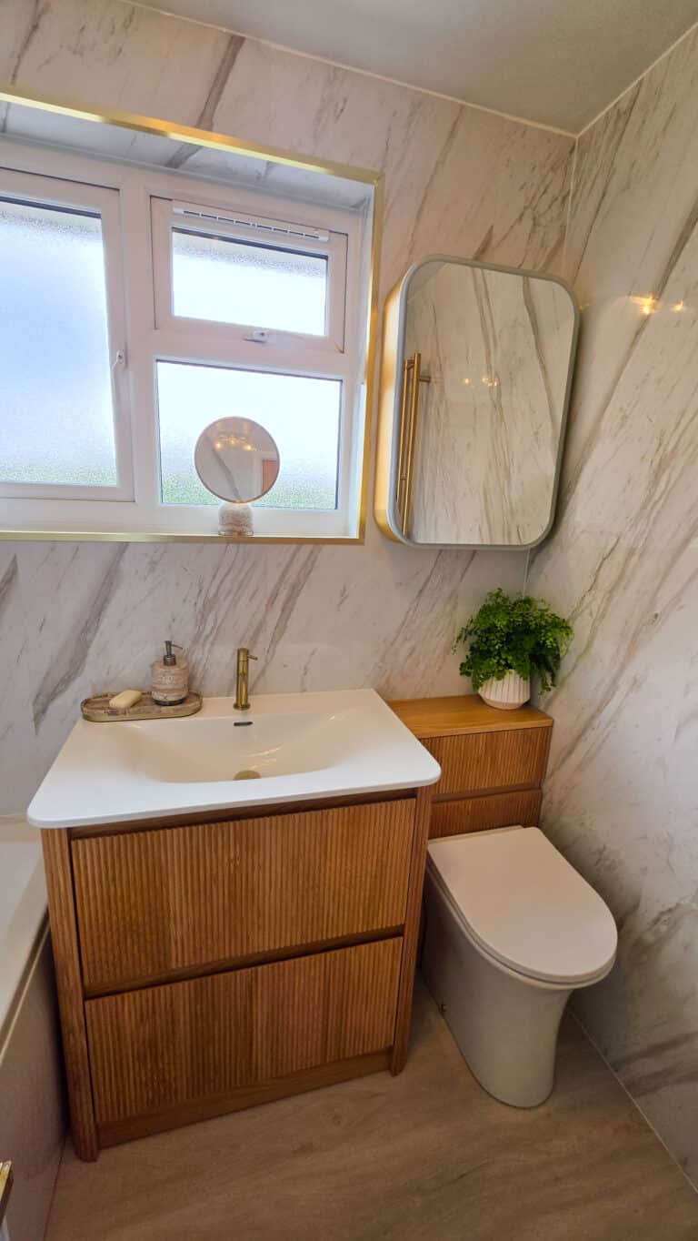 Small modern bathroom before and after transformation featuring marble walls, a wooden vanity with white basin, gold tap, wall mirrored cabinet, potted plant, and a frosted window providing natural light.