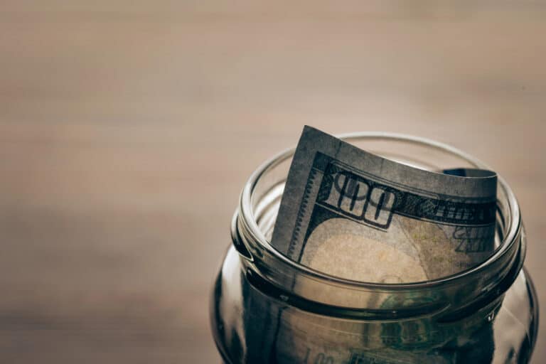A glass jar containing a folded one hundred dollar note, placed on a wooden surface with a blurred background—symbolising the importance of saving and investing after a market crash.