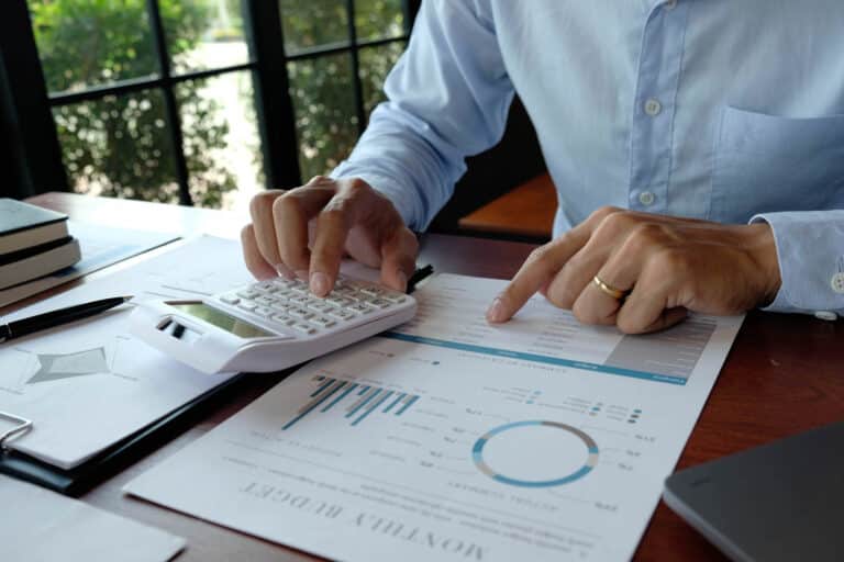 A person in a blue shirt uses a calculator and reviews financial documents with charts and graphs at a desk near a window, carefully considering strategies for investing after a market crash.