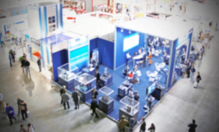 A blurred image of a trade show or expo, featuring people walking around various blue and white stands in a large indoor space, captures the dynamic atmosphere of co-exhibiting companies presenting their products and services side by side.