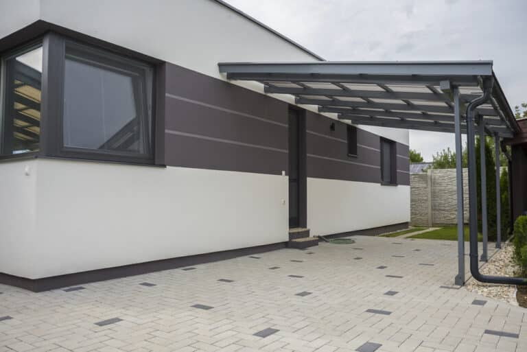 Modern house exterior with grey and white walls, large windows, a covered entrance with a metal roof, and a paved drive. Designed with self-care for busy parents in mind, the layout includes inviting outdoor areas where families can unwind amidst grass and greenery visible on the right side.