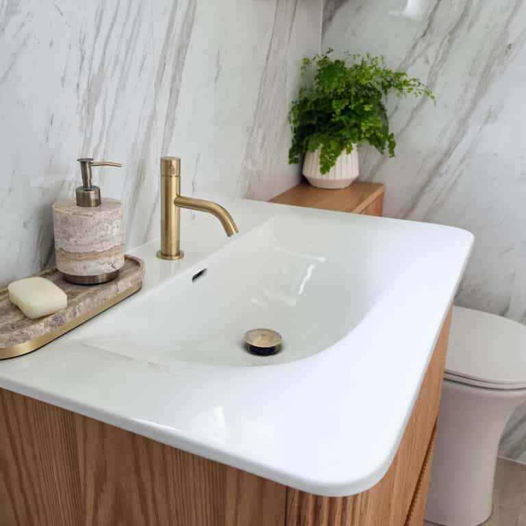 Modern bathroom basin with a gold tap, marble walls, a soap dispenser, and a tray with soap. A potted green plant sits on the wooden vanity unit, with a toilet partially visible in the background—showcasing attention to detail and refined design that rivals the expertise of top patio contractors in Beaverton.