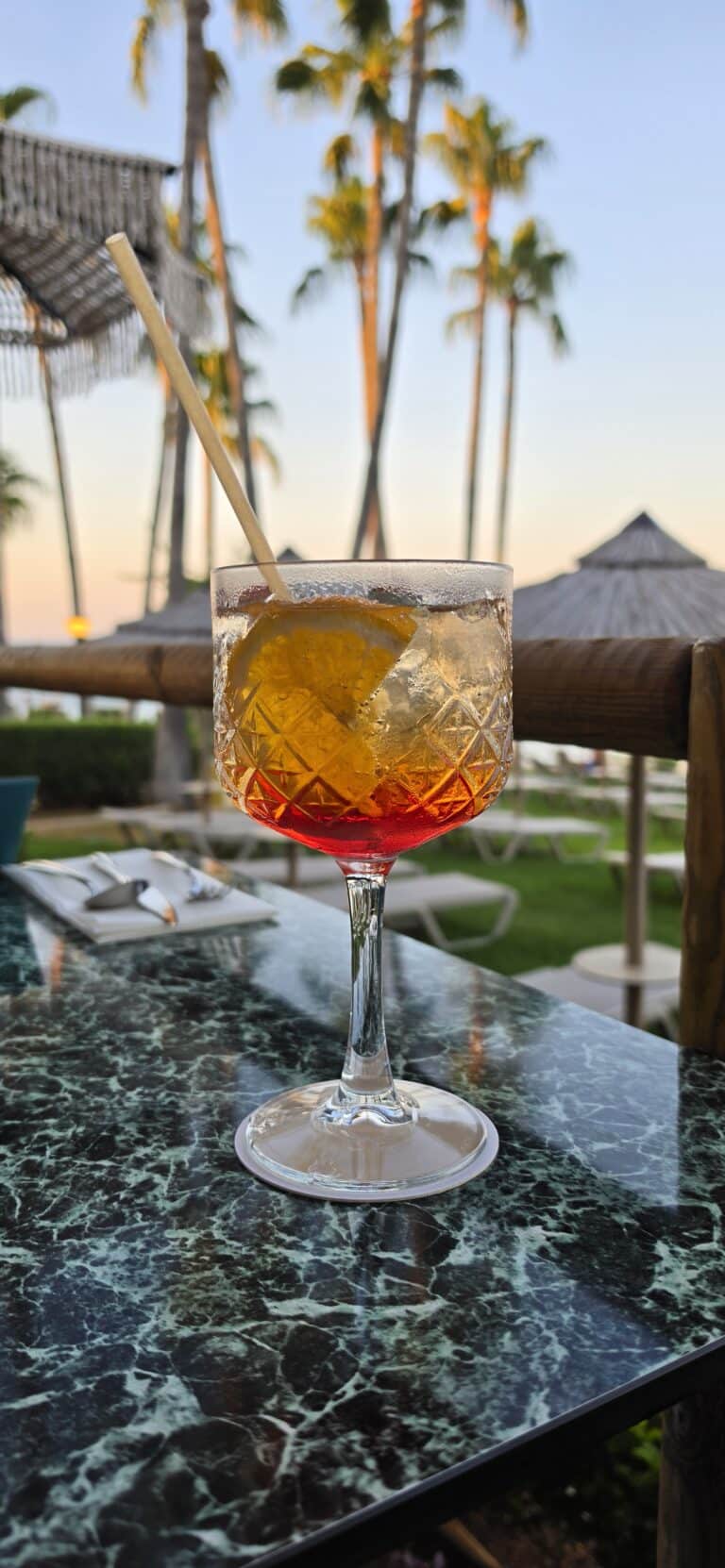 A cocktail with ice, a lemon slice, and a straw sits on a marble table outdoors. Palm trees and loungers are visible in the background at sunset, capturing the laid-back elegance of popular bars in Paros that define the island’s vibrant nightlife.
