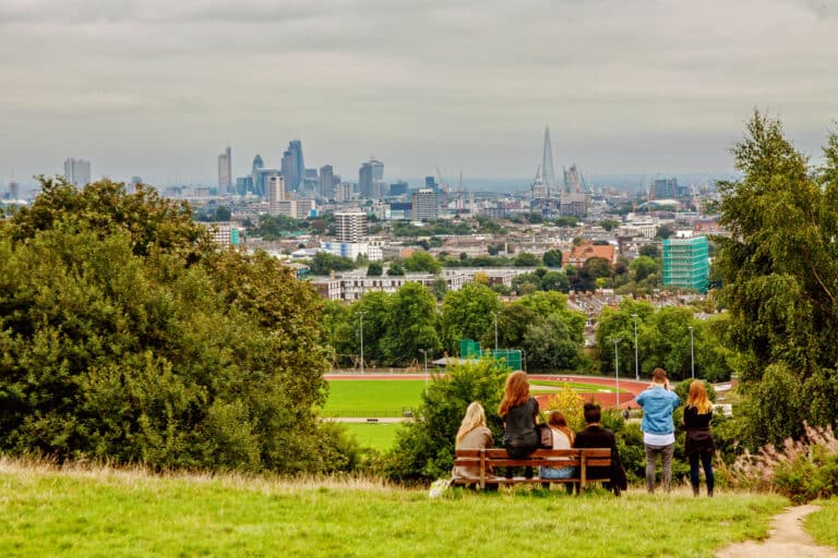 People sit and stand on a hill overlooking a city skyline with skyscrapers and a large park below, under a cloudy sky—capturing the spirit of a Family Day Out on Hampstead Heath.