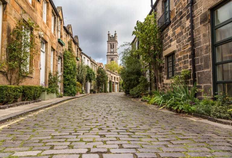 A narrow cobblestone street lined with stone buildings and greenery, with a tall church tower visible in the background under a cloudy sky—a timeless scene that reminds you of the importance of keeping your devices safe as you explore.
