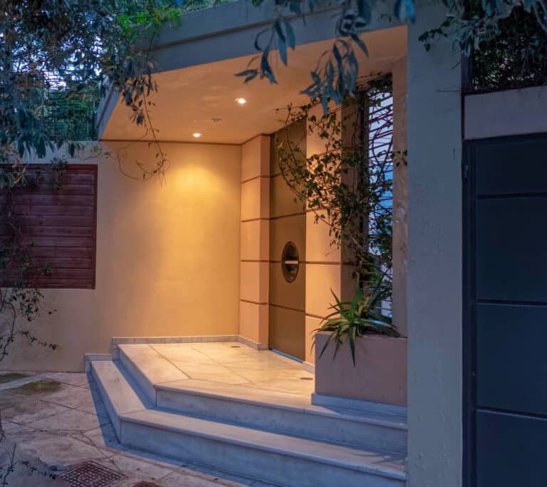 Modern house entrance with beige walls, a round windowed door, marble steps, built-in lights, and plants growing along the side—perfectly reflecting the elegance of a high-end property in Athens.