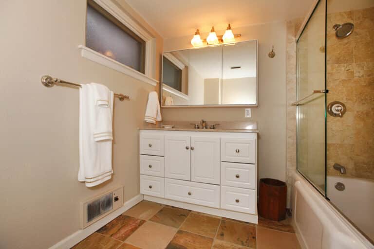 Modern bathroom with white vanity, mirror, and triple light fitting. Tiled floor and shower-bath combo with glass door. Two white towels hang on a metal rail next to the vanity. This space is equipped with a macerator pump for efficient wastewater management, seamlessly blending convenience with contemporary design.
