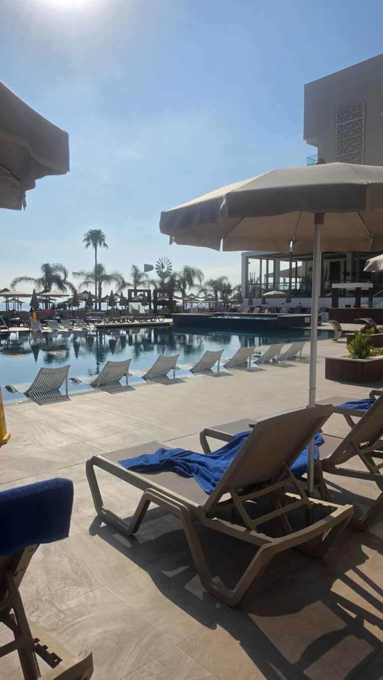 Lounge chairs with blue towels and umbrellas face a calm swimming pool, with palm trees and a modern building in the background under a bright, sunny sky—a tranquil setting that feels far removed from the hustle of managing your guest post marketplace.