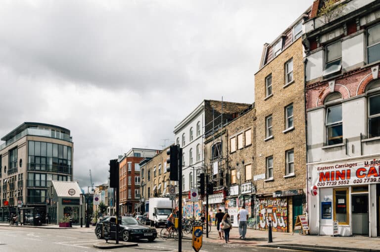 A city street scene with brick buildings, a Mini Cabs shop, graffiti on walls, pedestrians, streetlights, and cars. The sky is cloudy, and modern and older architecture line the street. Among the lively businesses is a bike repair in London shop, adding to the vibrant urban atmosphere.