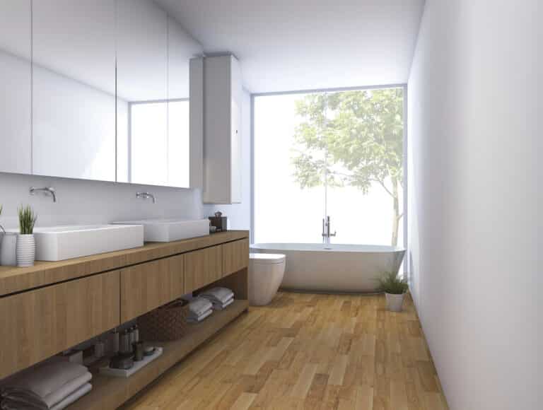 Modern bathroom with wooden floor, double sinks, open shelving with towels, a freestanding bath beside large sash bay windows, and green plants. Natural light enters through the sash bay windows, creating a bright, airy atmosphere.