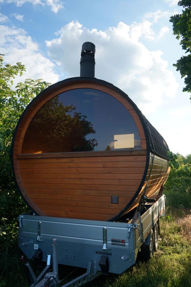 A barrel-shaped wooden sauna with a large window sits on a trailer in a grassy area, surrounded by green trees under a blue sky with scattered clouds—the perfect retreat to complement any stylish family home.