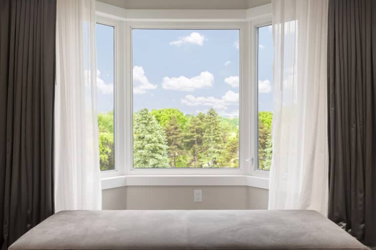 A large bay window with sheer curtains overlooks a bright, green landscape dotted with trees and a blue sky—an inspiring view for anyone considering the best outdoor saunas for small gardens. A grey cushioned bench sits below the window, inviting relaxation while you plan your perfect garden retreat.