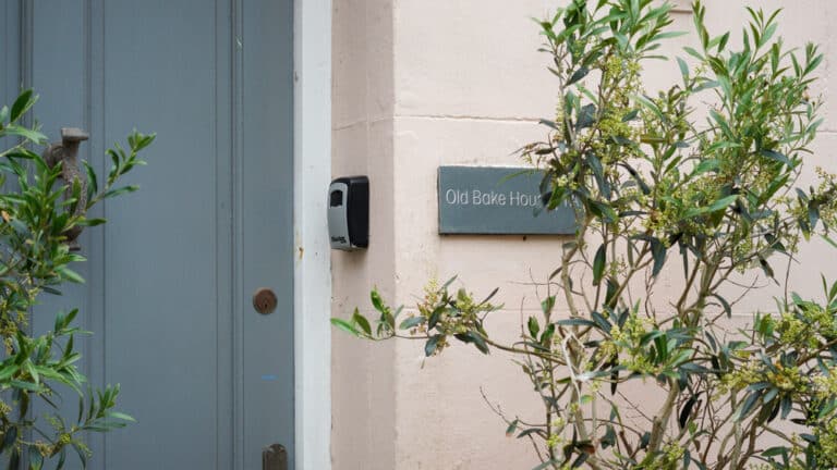 A pale pink wall with a sign reading Old Bake House beside a blue-grey door and a leafy green plant partially obscuring the view, hints at the building’s history—perhaps once housing essentials like an old grinder pump alongside its ovens.