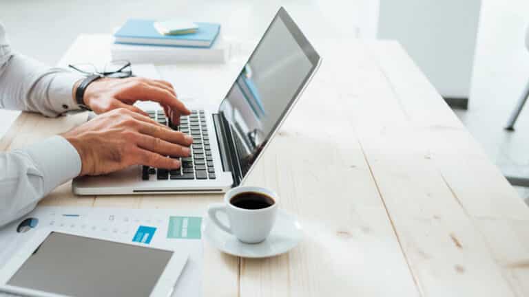 Person typing on a laptop at a wooden desk with documents, a tablet, notebooks, and a cup of coffee nearby in a bright office setting—researching Data Governance Tools and preparing to compare data governance companies for an upcoming project.