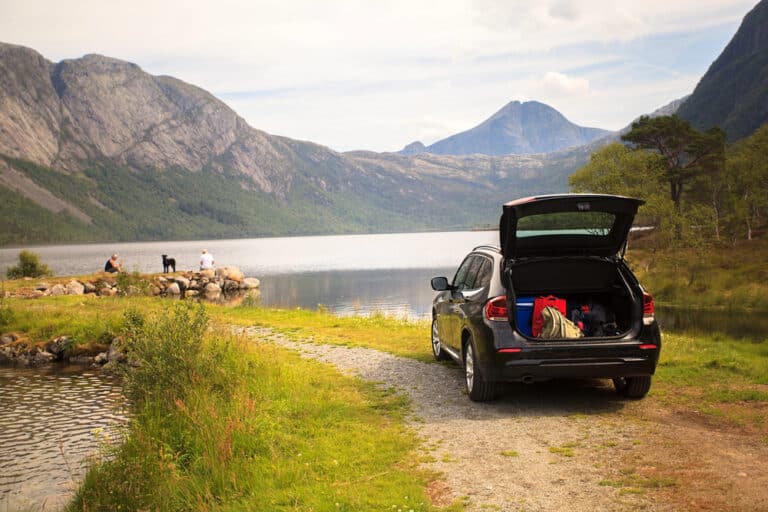A black SUV with its boot open, packed with gear, is parked by a lake surrounded by mountains—a perfect choice for family car hire. People and a dog sit by the water on rocks, enjoying the scenic, peaceful landscape.