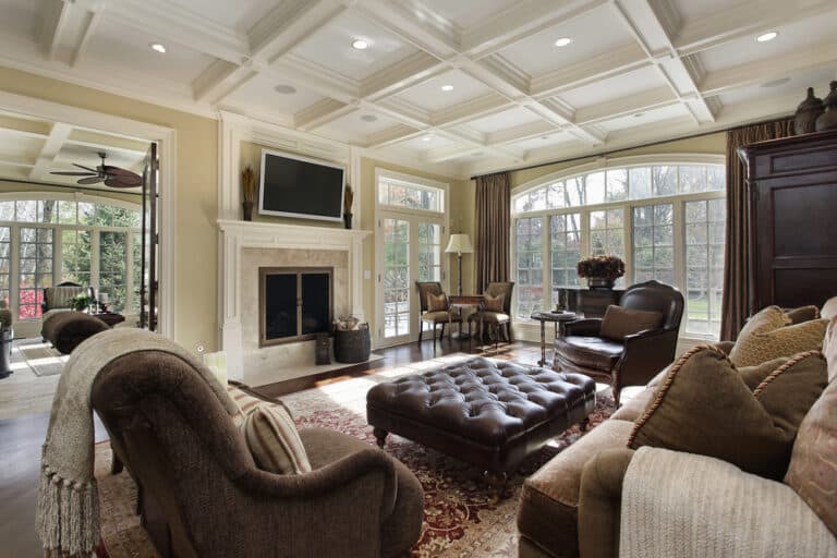 Elegant living room with coffered ceiling, fireplace, TV, large windows, plush sofas, armchairs, a tufted leather ottoman, and a rug. Thoughtfully designed as part of a Heritage Building Renovation, this inviting space is complemented by a sunlit sitting area visible through French doors.