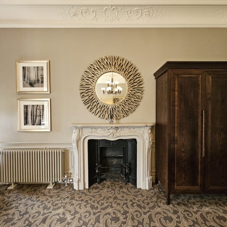 A beautifully restored heritage building renovation, this room features a decorative fireplace, an ornate mirror, two framed pictures, a radiator, patterned carpet, and a dark wooden wardrobe against beige walls with detailed cornicing.