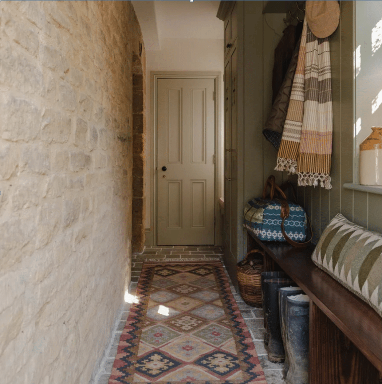 A corridor with a patterned rug, stone wall, green door, bench with cushions, boots underneath, hanging coats, hats, and a woven bag on a shelf—perfect for anyone building a modern home that effortlessly blends style and functionality.