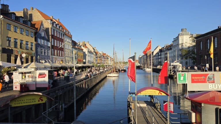 Nyhavn canal in Copenhagen, featuring colourful historic buildings, moored boats, and a canal tour boarding area with red flags where travellers might be asked to present a child travel consent form.