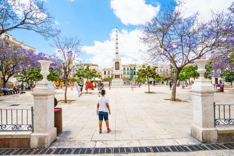 A child stands in a sunlit public square on their last day in Malaga, facing a tall stone obelisk surrounded by blooming purple jacaranda trees and several people.
