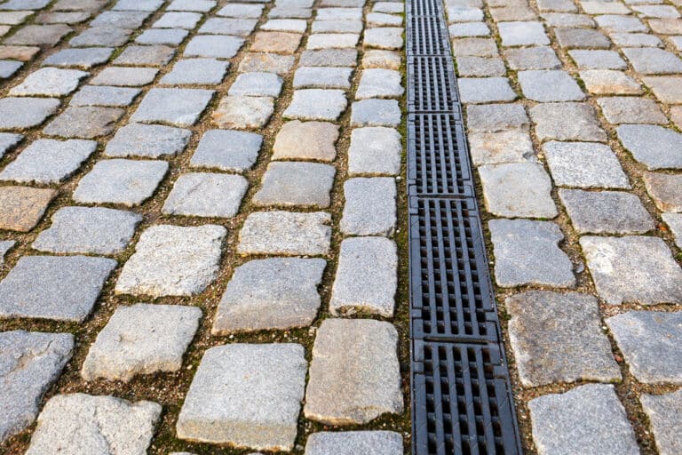 Cobblestone pavement with a black metal drainage grate running vertically through the centre—a key element for effective drainage and maintenance in commercial property. Moss and grass grow between some of the stones.