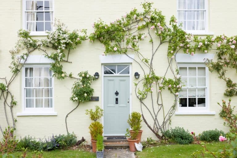 A light yellow cottage exterior with a pale green front door and climbing pink rose vines, reflecting the cosy, secure environment sought in security tips for new parents.