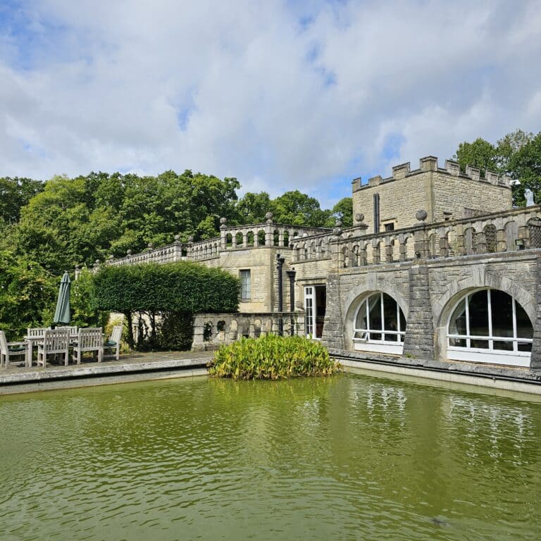 A stone building with castle-like features stands behind a pond with green water and plants, surrounded by trees and outdoor seating—a serene spot reflecting strong data governance foundations.