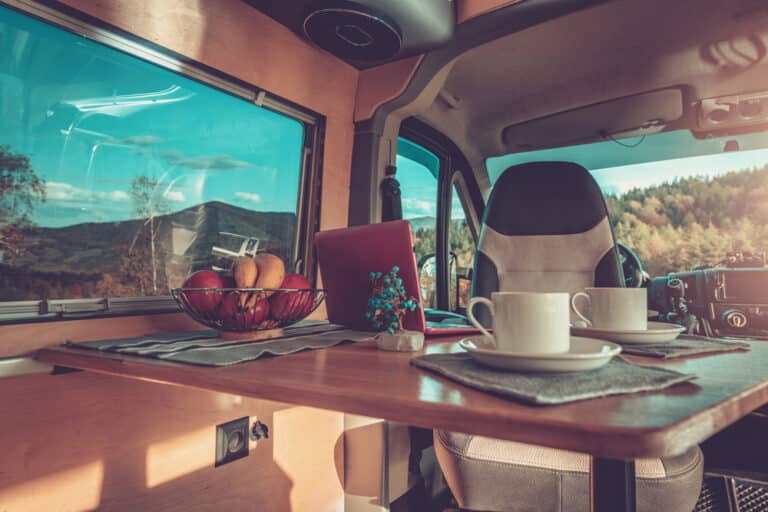 Interior of a camper van featuring one of our camper conversion kits, with a wooden table holding two coffee cups, a laptop, and a bowl of fruit, overlooking a mountain landscape through the window.